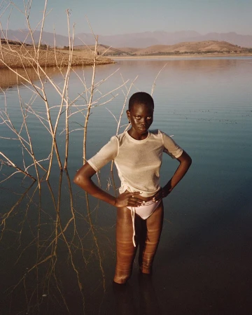Elegant woman stands waist-deep in water, posing with hands on hips. Background features lake, distant mountains, and sparse trees.