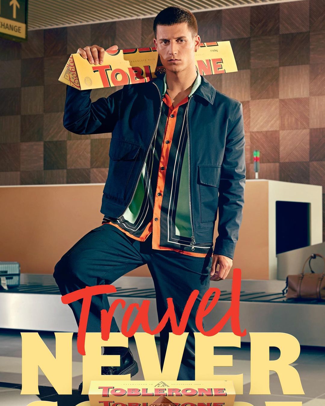 Adult male model in travel themed fashion editorial, holding Toblerone chocolate box at airport baggage claim with patterned background.