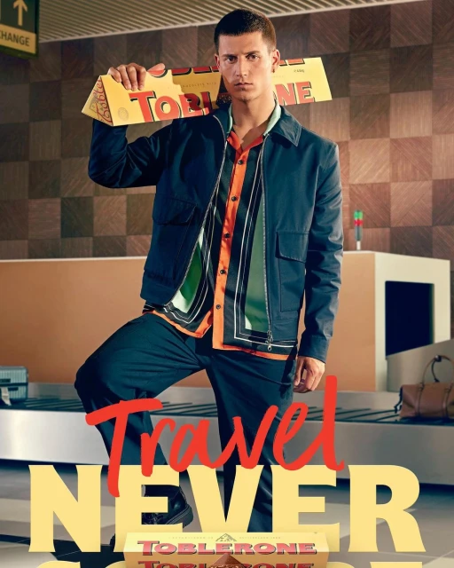 Adult male model in travel themed fashion editorial, holding Toblerone chocolate box at airport baggage claim with patterned background.