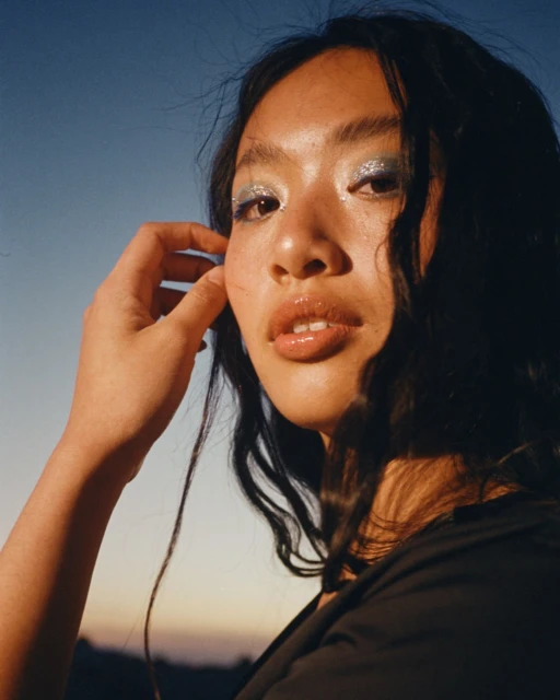 Close-up portrait of a young Asian woman with glitter eyeshadow, glossy lips, and black top against a blue and orange sky at dusk.