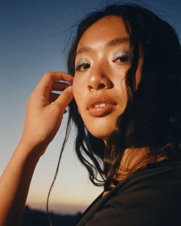 Close-up portrait of a young Asian woman with glitter eyeshadow, glossy lips, and black top against a blue and orange sky at dusk.