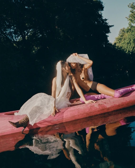 Two women in a canoe on a body of water surrounded by trees, one in a white gown, the other in lingerie and magenta boots.