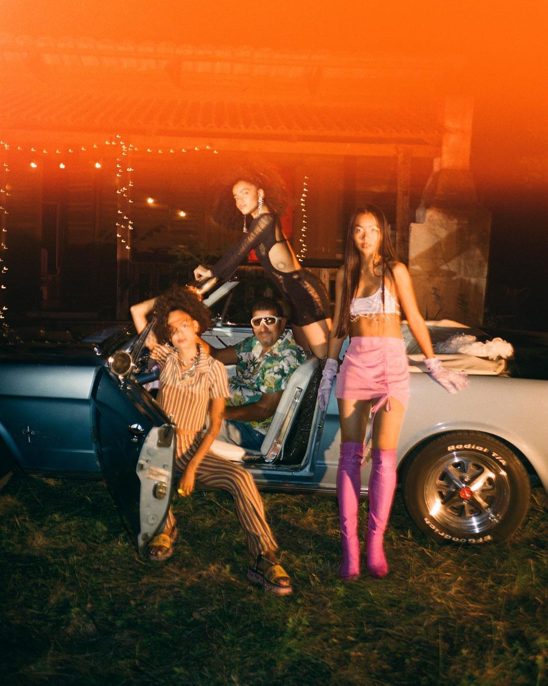 Four people pose in front of a classic blue convertible car at night, with a house illuminated with string lights in background.