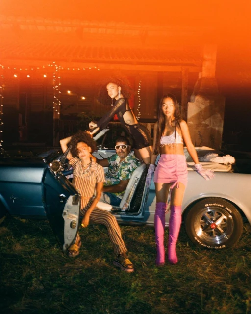 Four people pose in front of a classic blue convertible car at night, with a house illuminated with string lights in background.