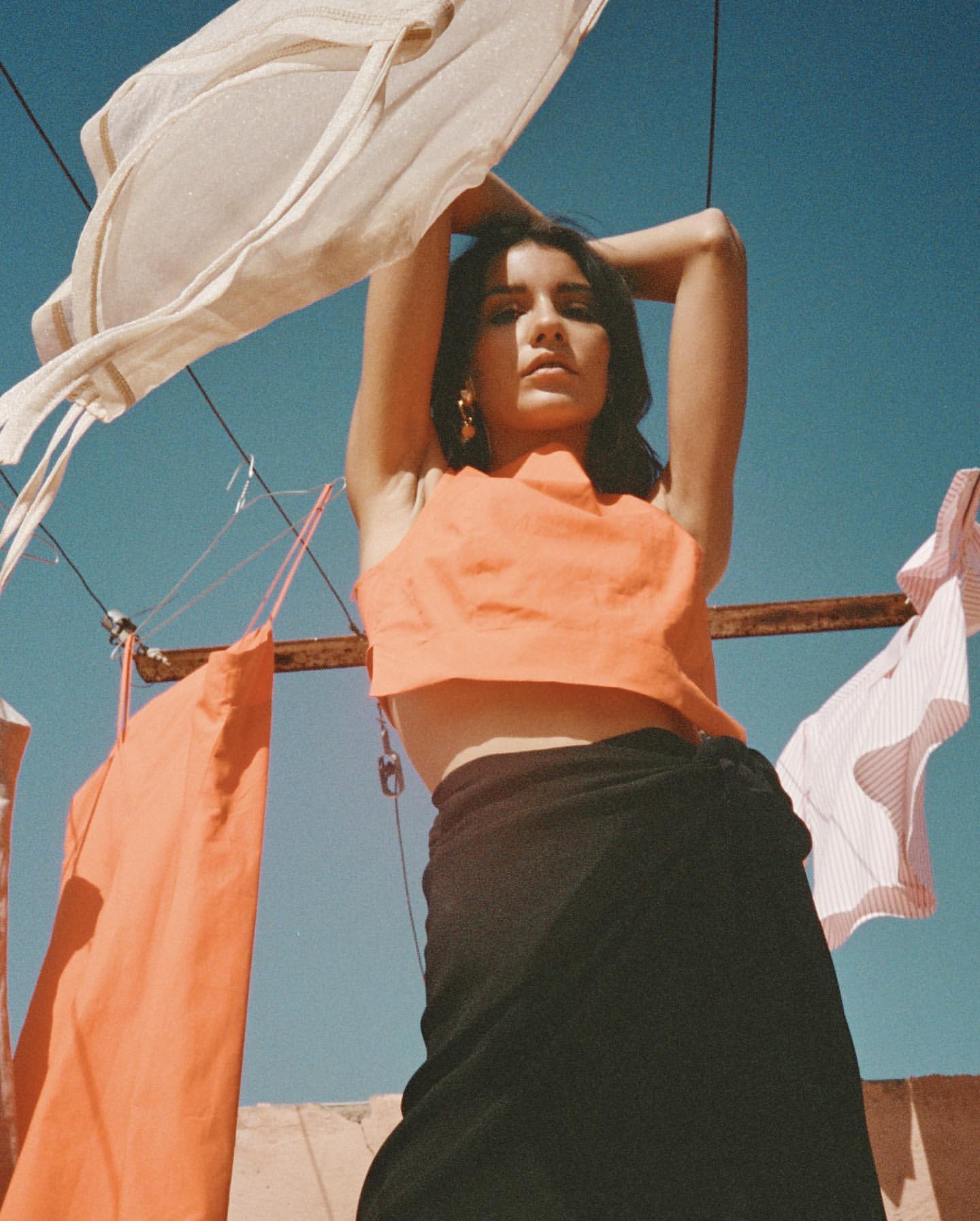 Woman posing under clothesline with flowing laundry. She wears an orange top and black skirt against a bright blue sky backdrop.