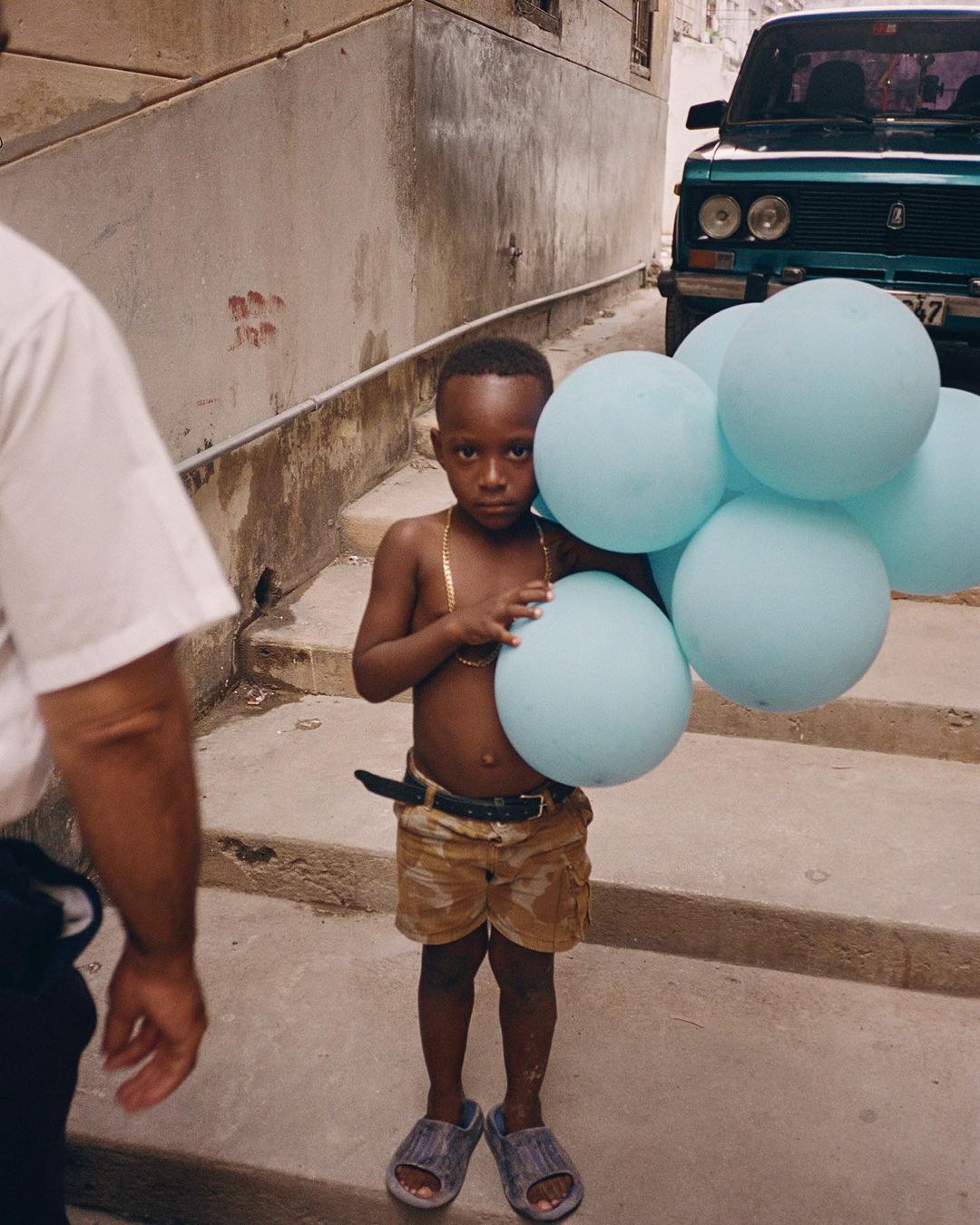 A young black child stands on concrete steps outdoors holding a bunch of pale blue balloons, wearing shorts, sandals and a gold chain.