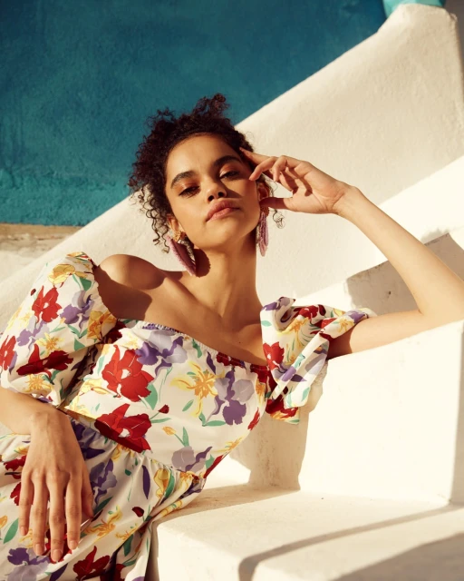 Attractive curly-haired woman in a floral dress reclines on white stairs, posing in sunlight against a blue wall in an outdoor setting.