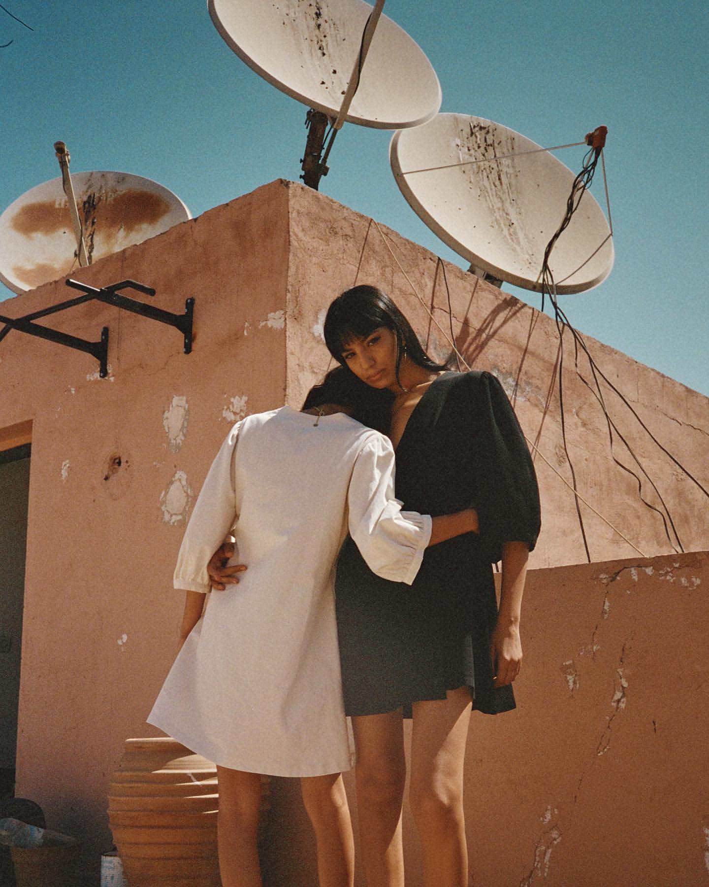 Two women embrace on a rooftop with satellite dishes against a clear sky, showcasing a blend of fashion and urban scenery.