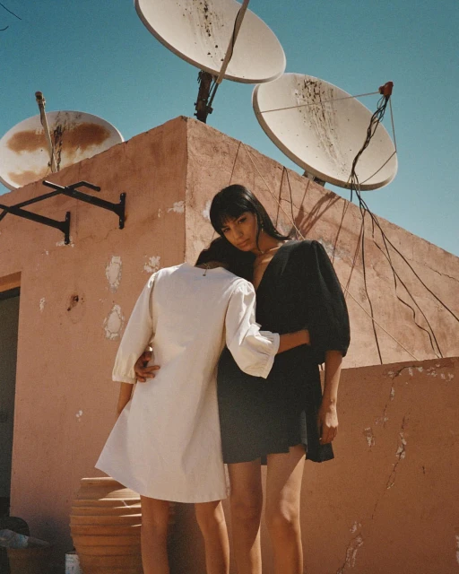 Two women embrace on a rooftop with satellite dishes against a clear sky, showcasing a blend of fashion and urban scenery.