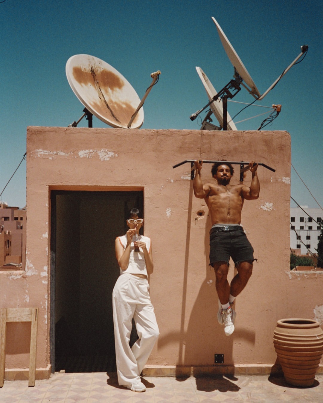 A man does pull-ups on a roof next to a woman in white pants; satellite dishes and clear blue sky overhead in an outdoor setting.