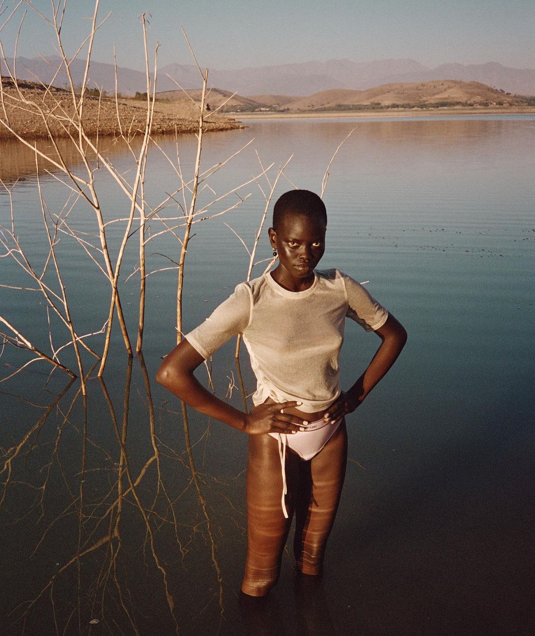 Woman standing in water by dead tree on lake with mountain background in outdoor shot, looking directly at the camera.