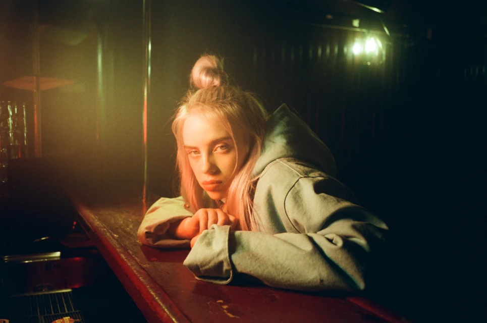 Blonde woman with hair in a bun, wearing a hooded jacket, leans against a red bar in a dimly lit room with golden light.