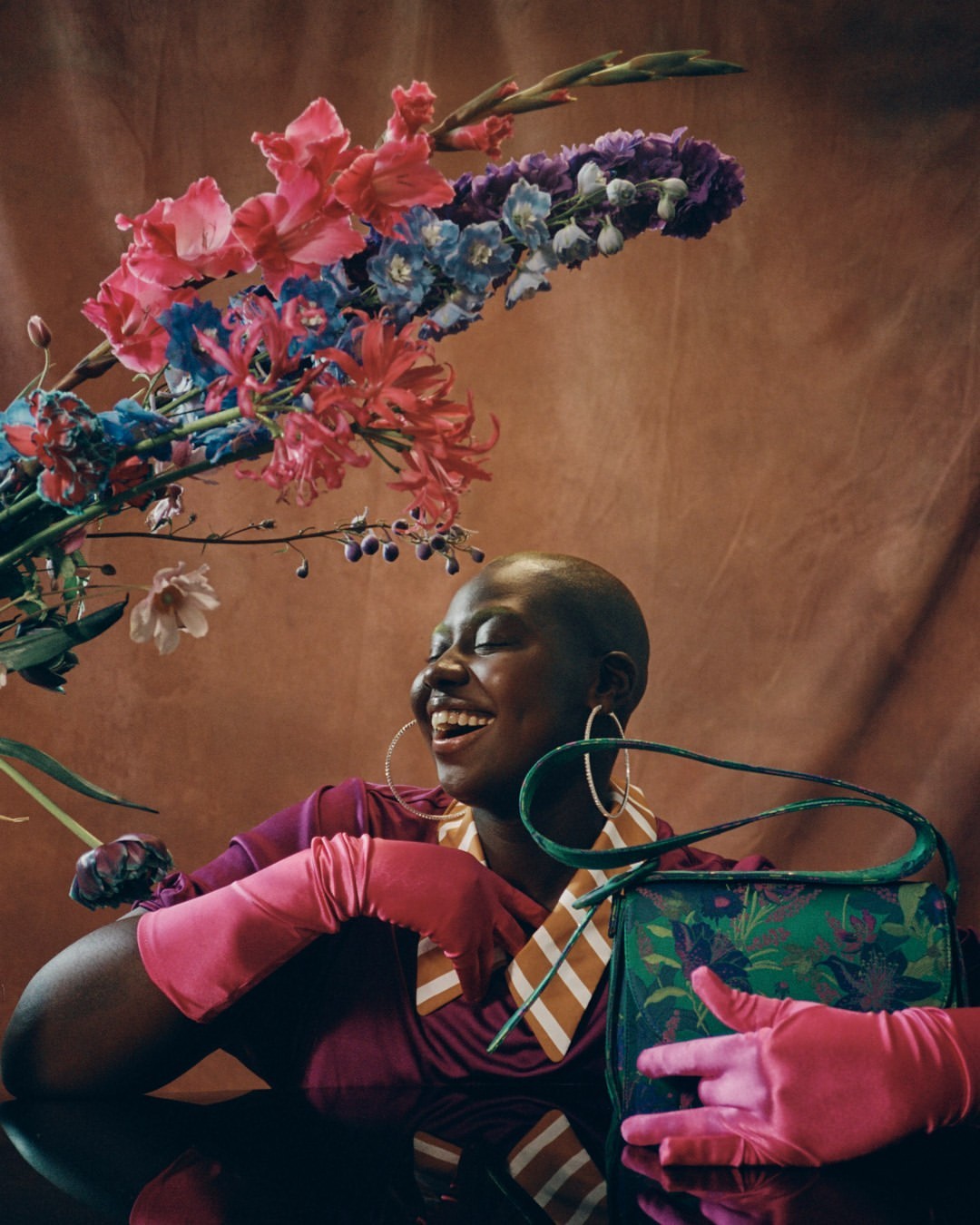 Smiling Black woman in pink gloves next to a purse and flowers, posing against a brown backdrop in a studio setting. 