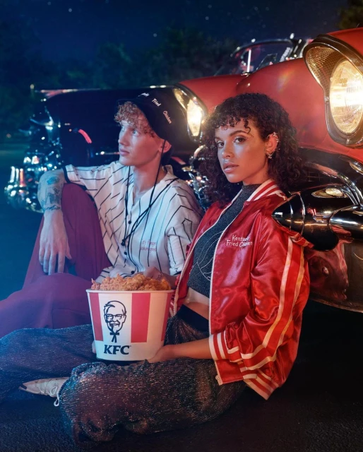 Two young adults sit beside a vintage car, sharing a bucket of KFC fried chicken at night, illuminated by cool-toned and red lighting.