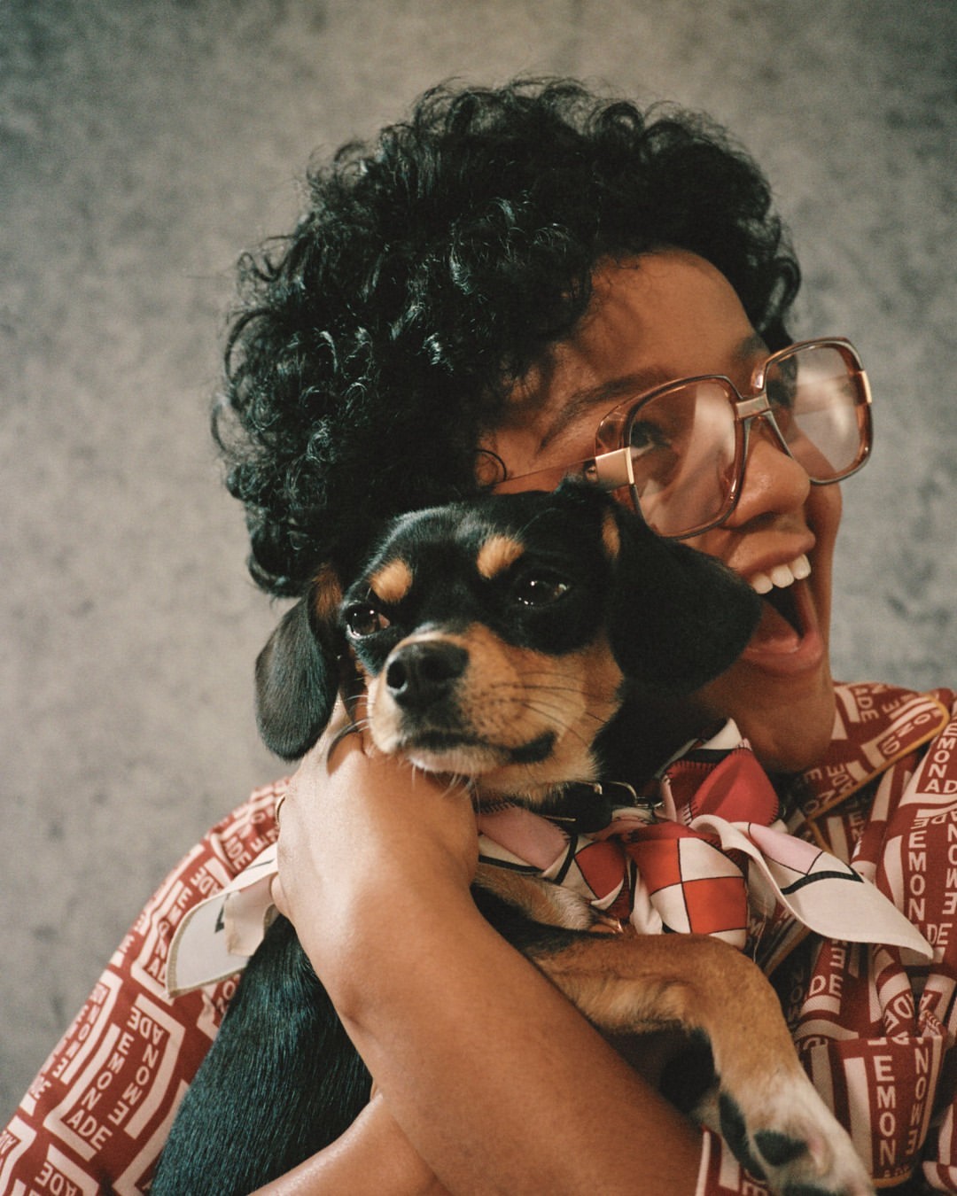 Smiling woman wearing glasses holds a small dog with a patterned scarf indoors, showcasing a casual and joyful moment between person and pet.