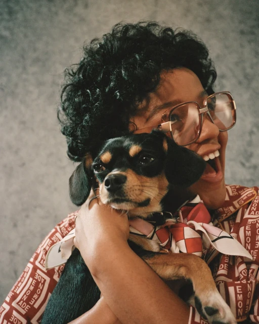 Smiling woman wearing glasses holds a small dog with a patterned scarf indoors, showcasing a casual and joyful moment between person and pet.