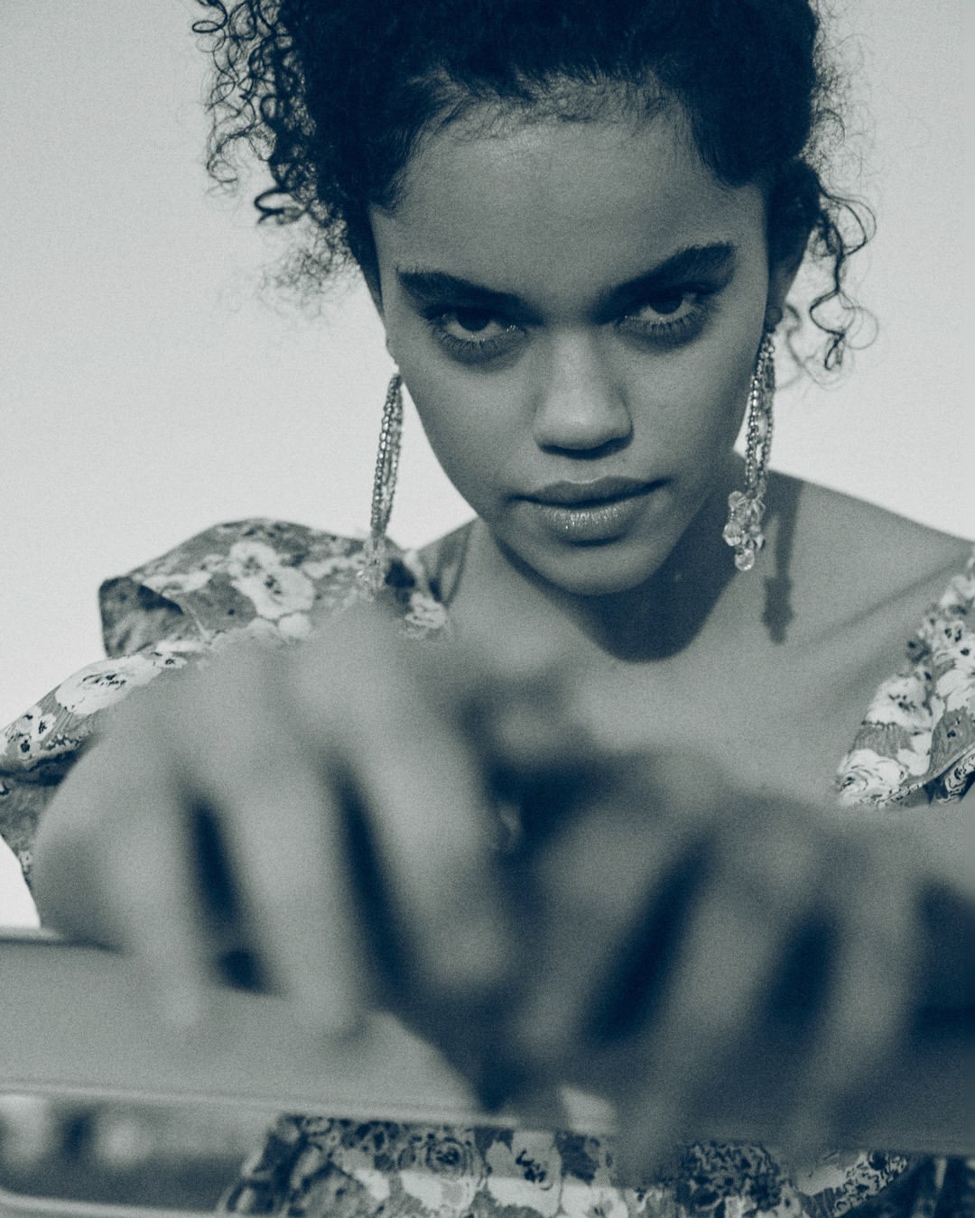 Portrait of a young woman with curly hair wearing large earrings and a floral top, gazing intensely at the camera in soft, diffused light.