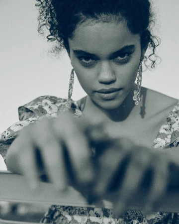 Portrait of a young woman with curly hair wearing large earrings and a floral top, gazing intensely at the camera in soft, diffused light.