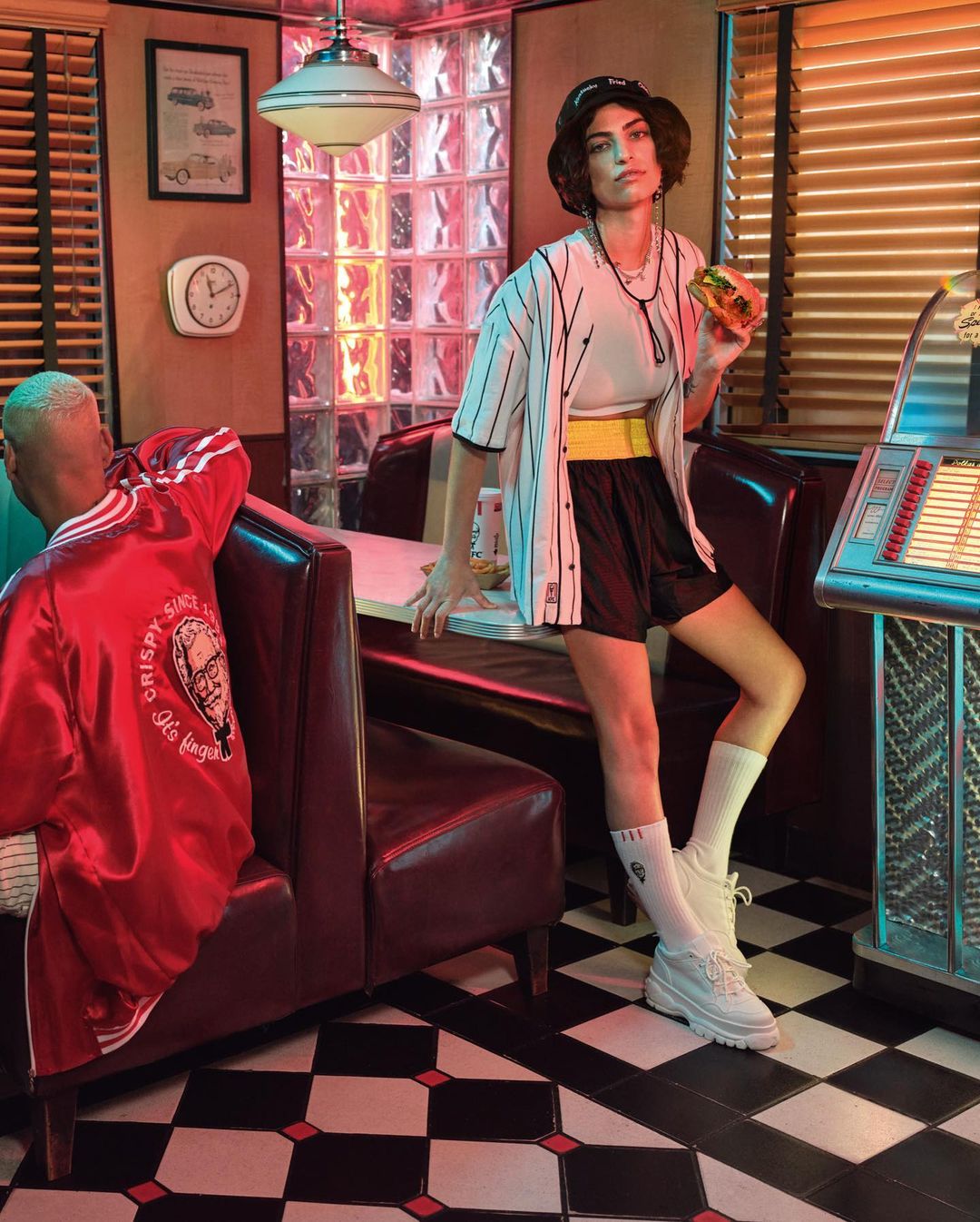 A woman poses in a retro diner with a burger, wearing a baseball jersey and hat, seated next to a vintage jukebox and booth seating.