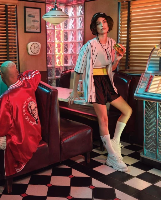A woman poses in a retro diner with a burger, wearing a baseball jersey and hat, seated next to a vintage jukebox and booth seating.