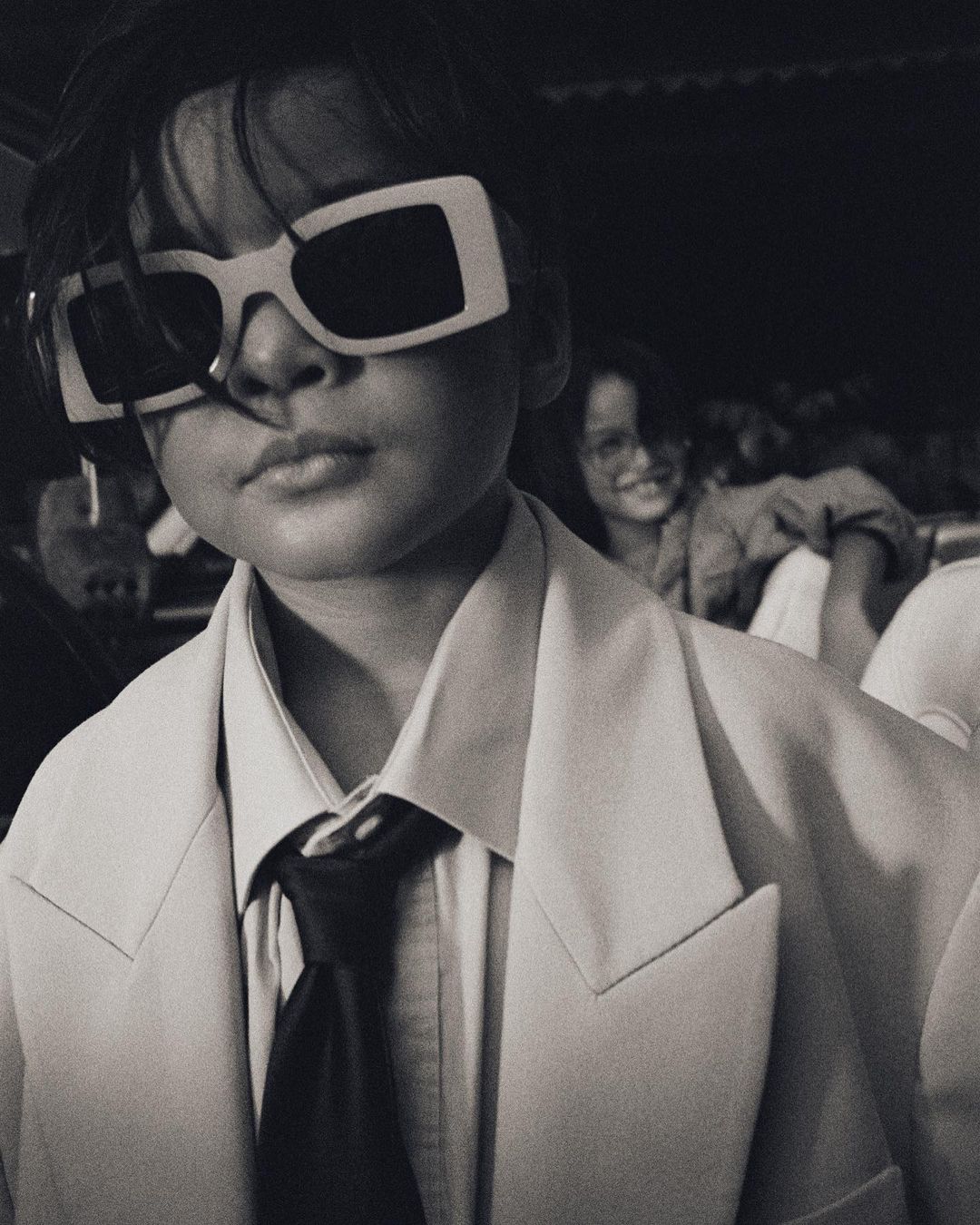 Close-up of a child wearing oversized white frame sunglasses, a pale blazer, and a black tie, photographed by Manu Fauque.