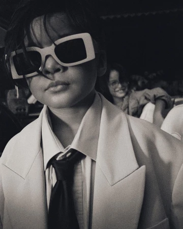 Close-up of a child wearing oversized white frame sunglasses, a pale blazer, and a black tie, photographed by Manu Fauque.