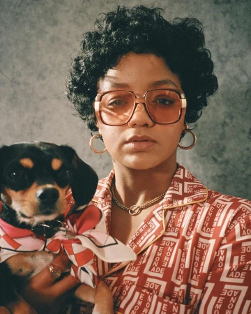 Portrait of a woman wearing tinted square glasses and a patterned red top holding a small black and tan dog with a matching scarf