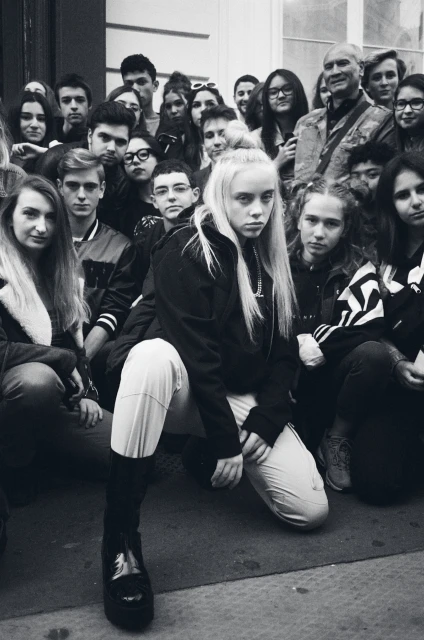 Black and white photo of a blonde woman kneeling in front of a large group of people in an urban setting, captured in an eye-level shot.