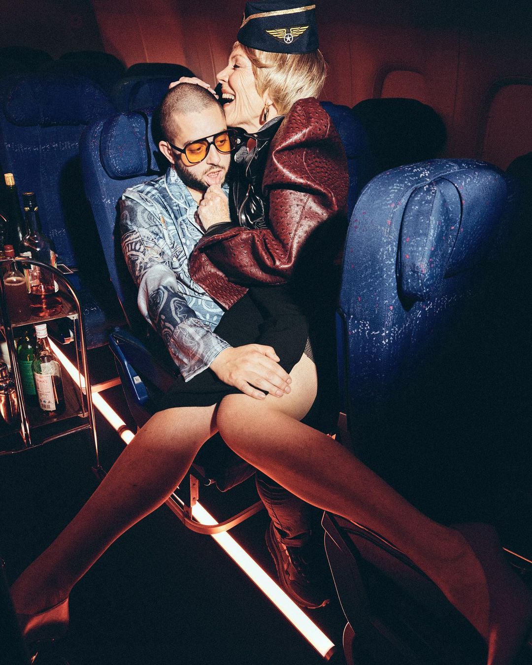 Woman in flight attendant uniform embraces man with glasses on airplane, backlit by lighting in cabin with drinks on rolling cart.