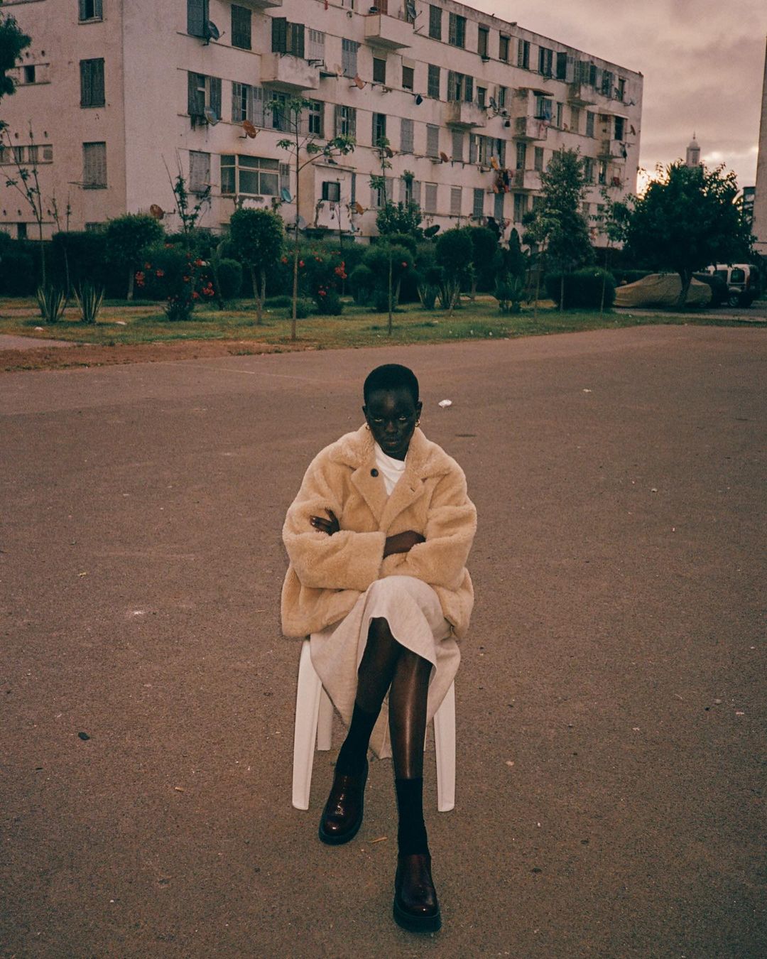 A young Black female model sits on a white chair wearing a beige coat and dark shoes in an urban outdoor setting with buildings.