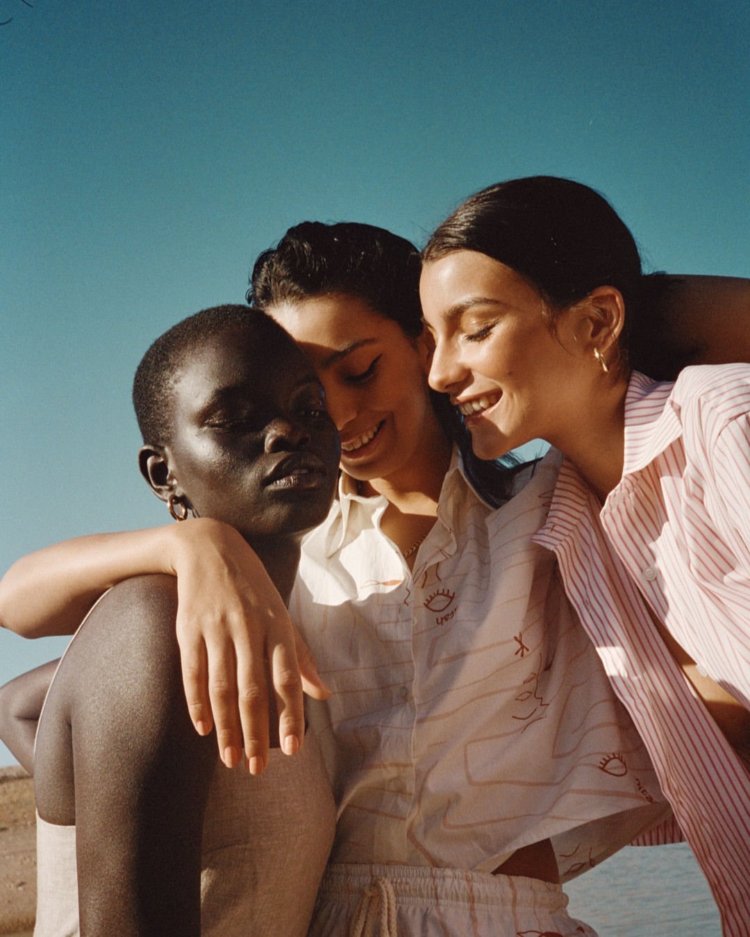 Three diverse young adult women embrace outdoors under clear sky, wearing casual patterned shirts in a close-up medium shot.