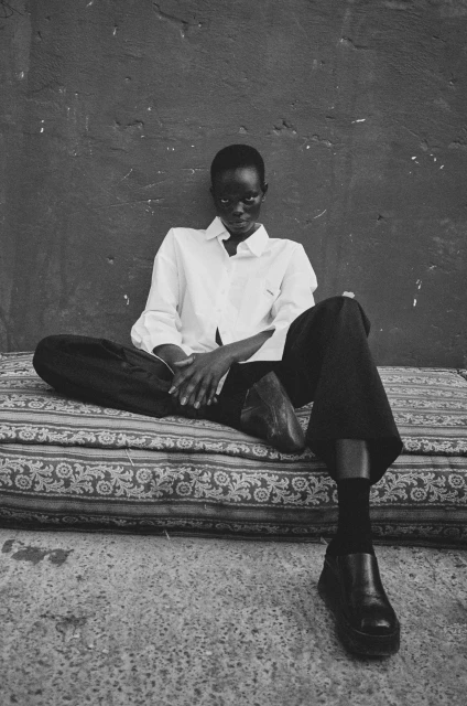 Black and white portrait of a short-haired adult woman sitting on an patterned cushion wearing a white shirt and dark pants and shoes.