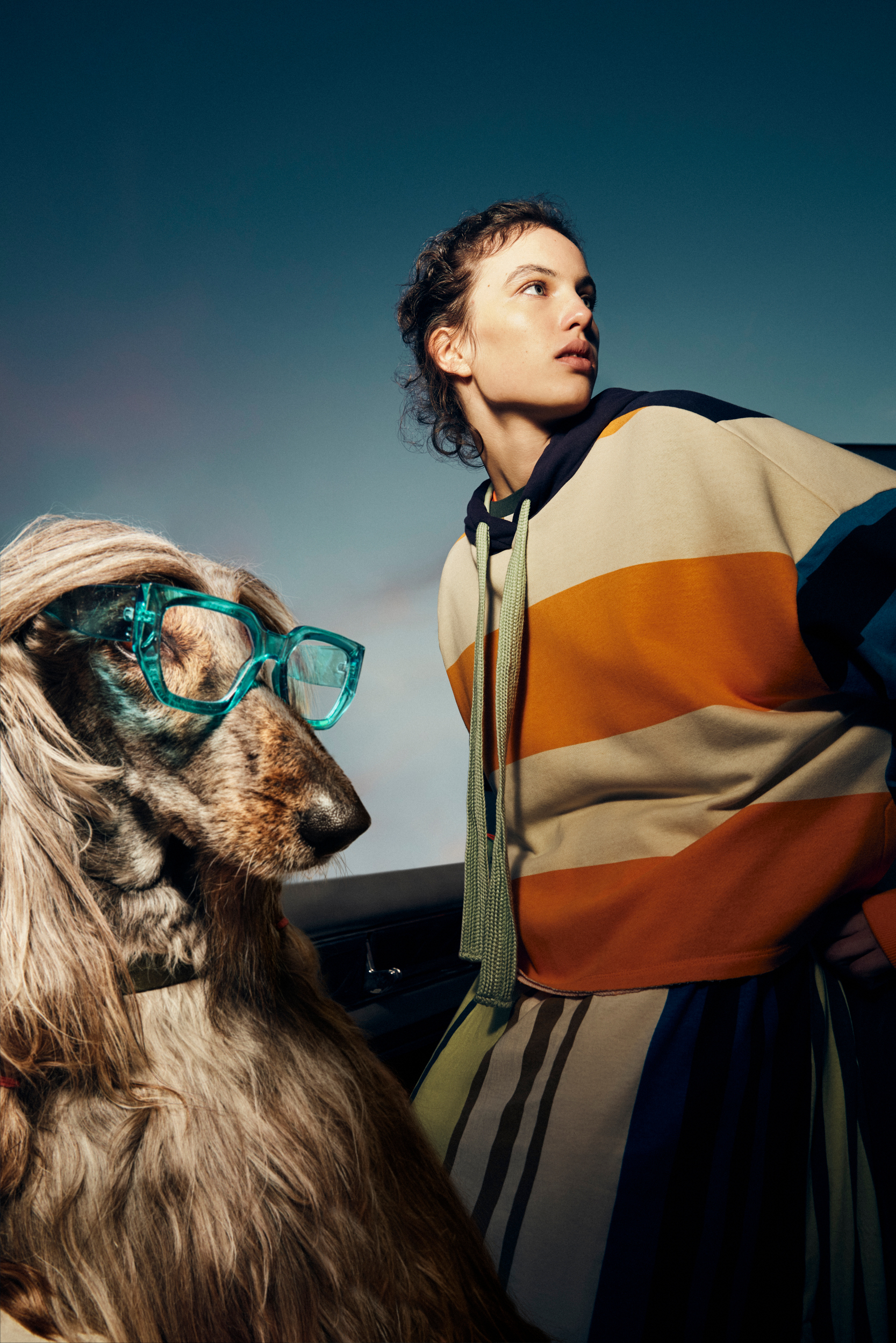 A young woman in a striped hoodie and a dog wearing teal eyeglasses sit side by side, looking off into the distance against a blue gradient backdrop.