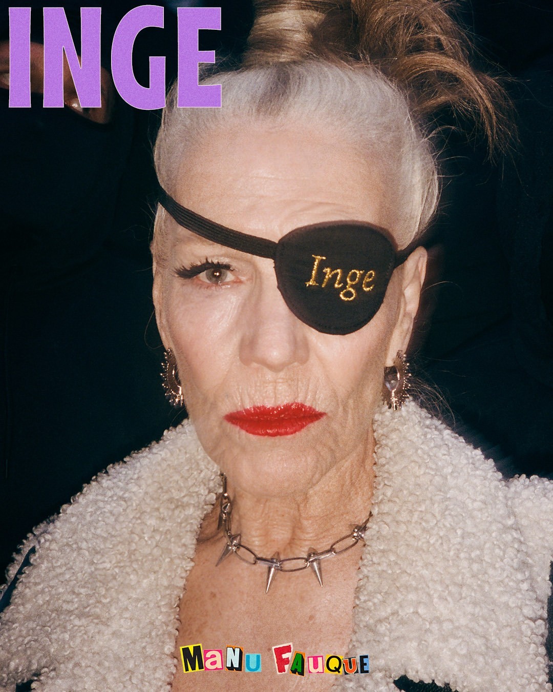 Elderly woman with an eyepatch and bold red lipstick wearing a spiked collar, a fuzzy coat and earrings, in a close-up portrait.