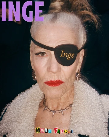 Elderly woman with an eyepatch and bold red lipstick wearing a spiked collar, a fuzzy coat and earrings, in a close-up portrait.