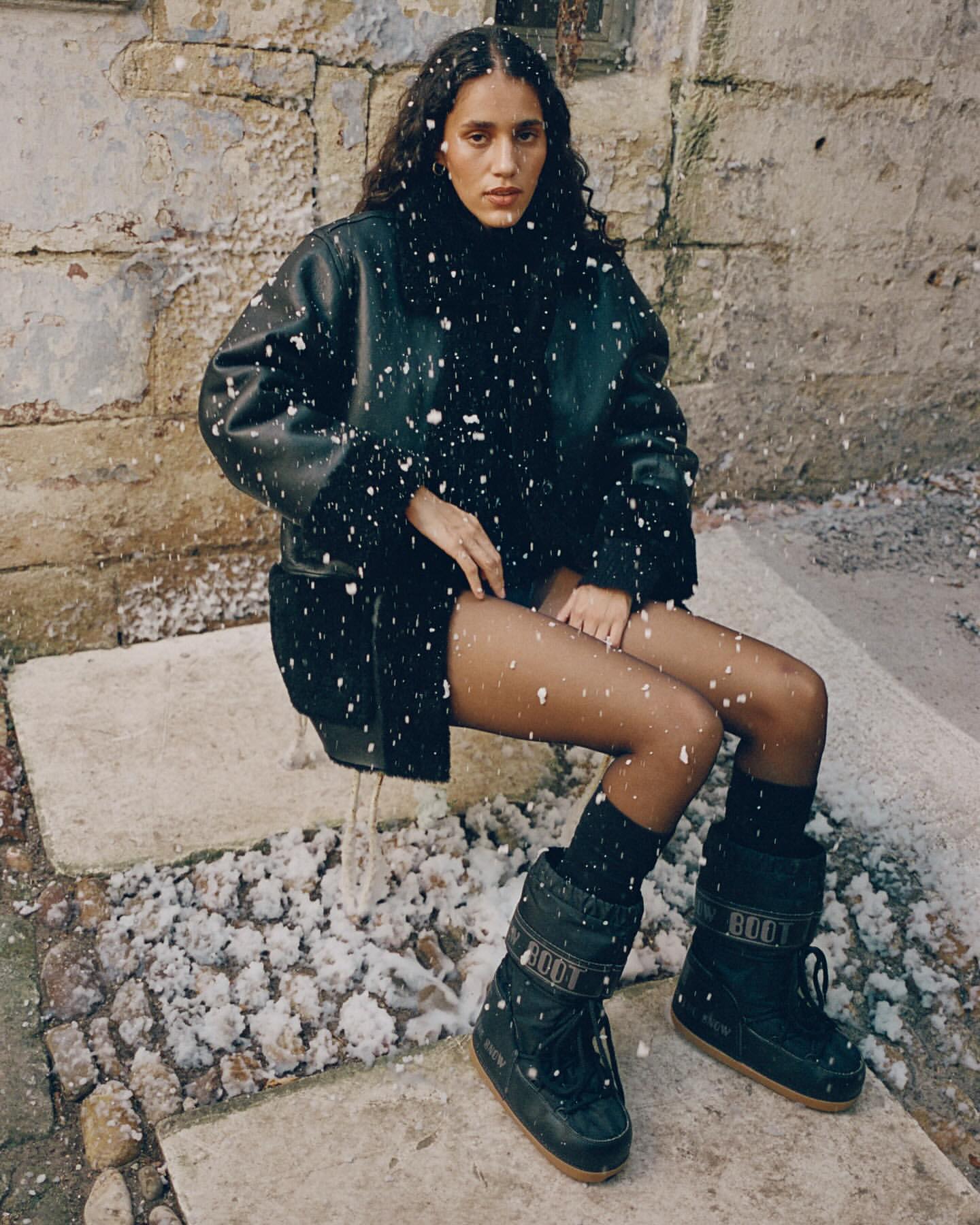 Young woman with curly dark hair, wearing black boots and a matching jacket, sits outside against a neutral wall in falling snow.
