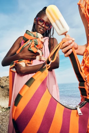Two models on the beach, one holding a small dog dressed in matching fabric, the other with a popsicle and striped bag.