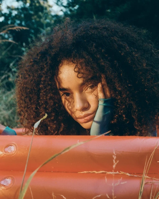 Young woman with afro resting head on hand, orange pool in an outdoor setting with tall grass. Soft natural light portrait.