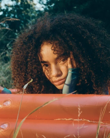 Young woman with afro resting head on hand, orange pool in an outdoor setting with tall grass. Soft natural light portrait.