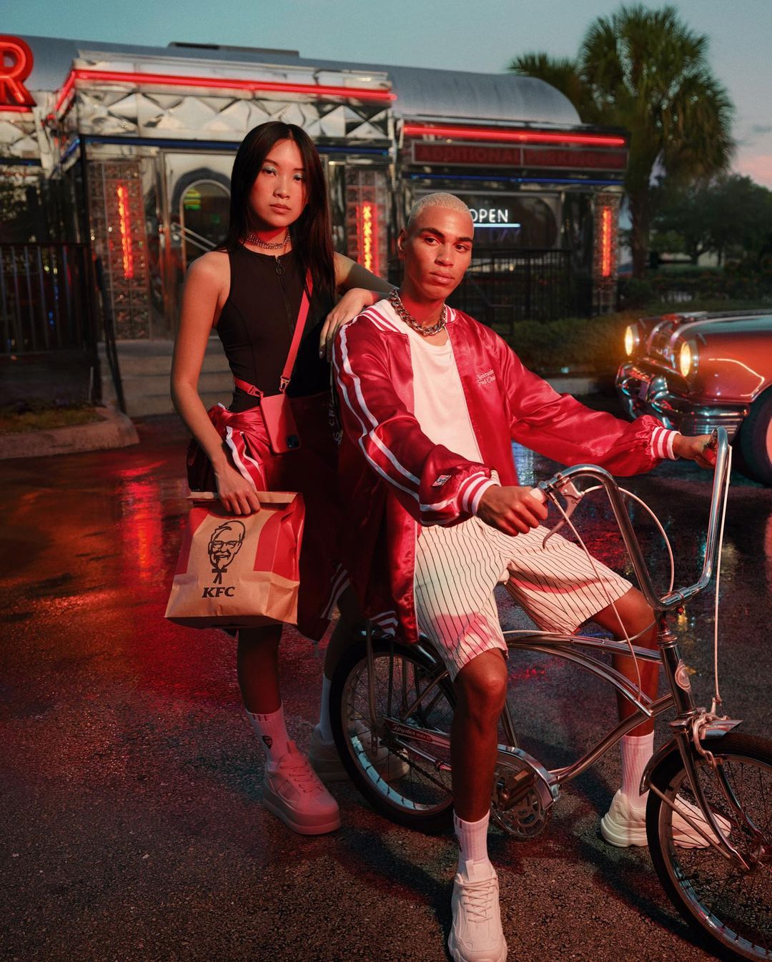 Two fashion models pose at dusk near a retro diner with a paper bag from KFC. A red vintage car is parked nearby on the wet street.