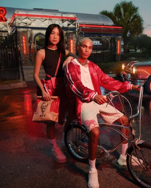 Two fashion models pose at dusk near a retro diner with a paper bag from KFC. A red vintage car is parked nearby on the wet street.