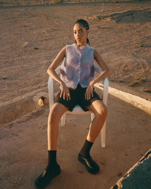 Young woman poses on a white chair outdoors, wearing a light purple sweater vest, dark shorts, black socks, and black platform shoes.