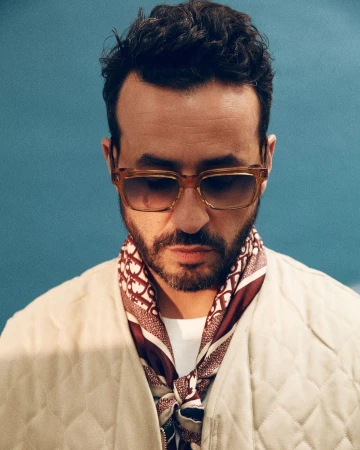 Man wearing amber-tinted sunglasses, patterned silk scarf, and quilted cream jacket against a textured blue background
