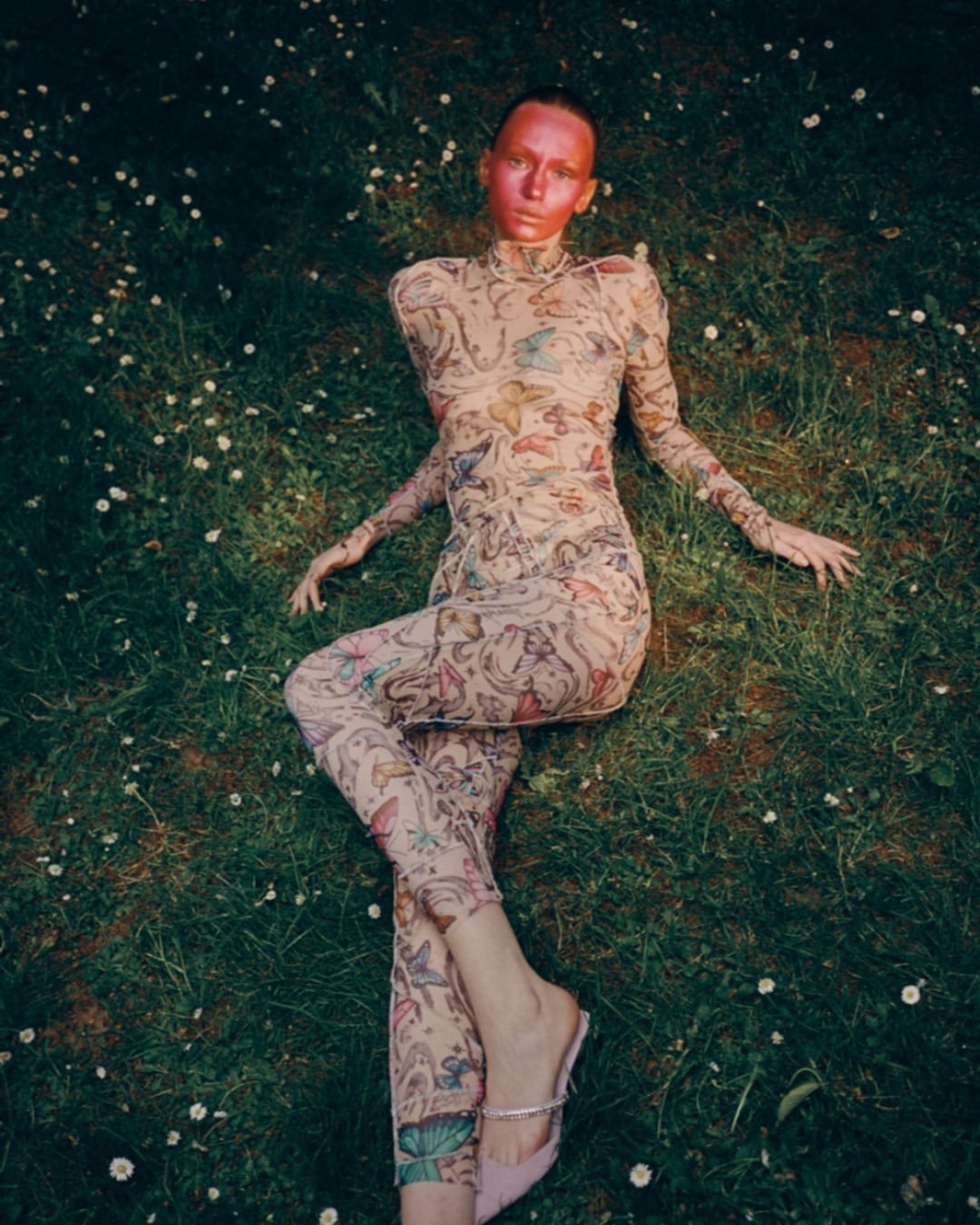 Woman with red face paint lies in grassy field wearing butterfly-print bodysuit and pink heels, captured in a high-angle outdoor shot.