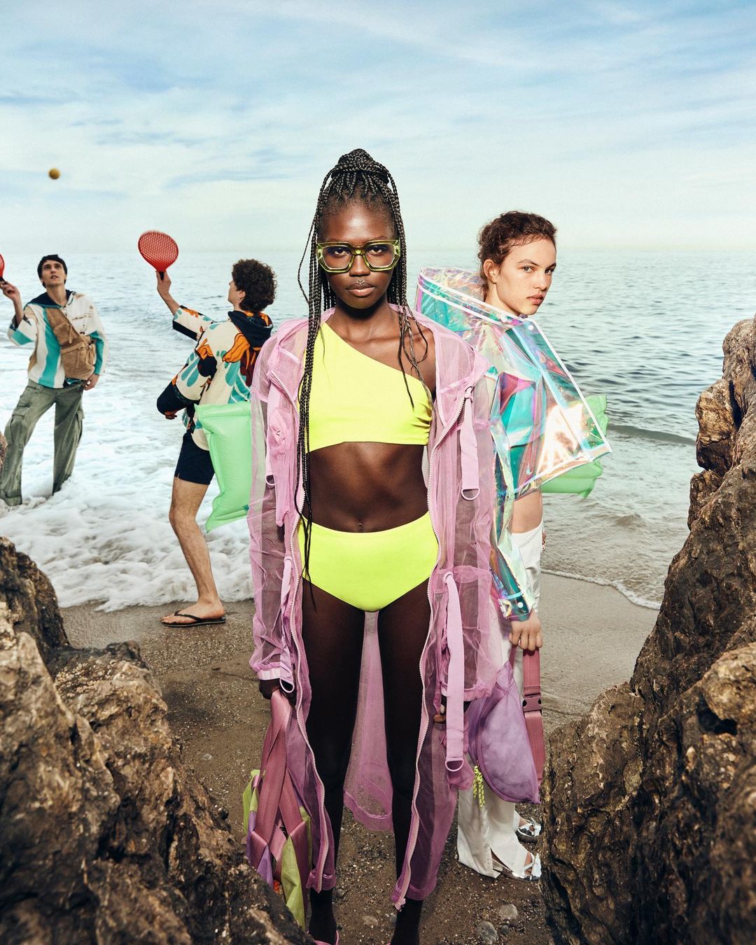 Two fashion models pose on a beach with other models playing in the background. The main model wears a yellow bikini and a pink coat.