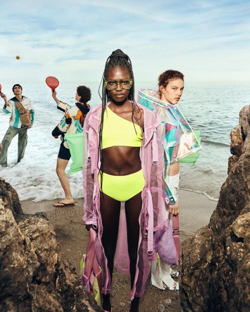 Two fashion models pose on a beach with other models playing in the background. The main model wears a yellow bikini and a pink coat.