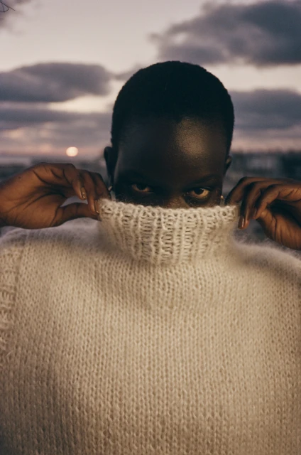 Close-up studio shot of a Black woman wearing a white turtleneck sweater, holding the collar up, with a dusk sky background.