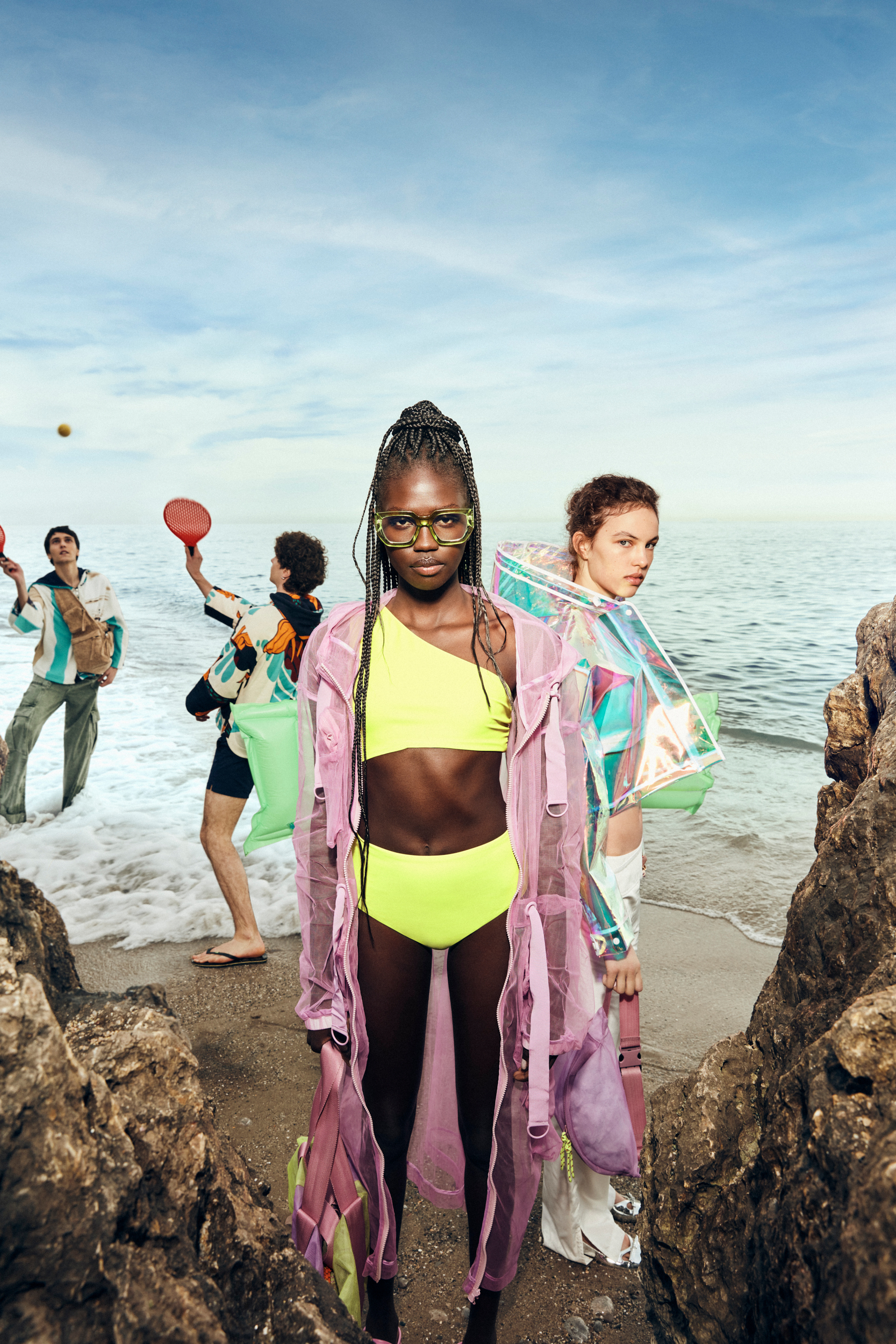 Editorial fashion shoot on a beach with models in colorful, transparent clothing, set against a backdrop of the ocean and sky.