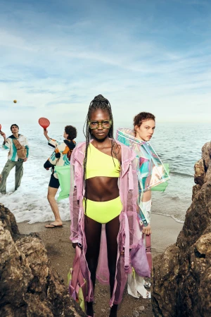 Editorial fashion shoot on a beach with models in colorful, transparent clothing, set against a backdrop of the ocean and sky.