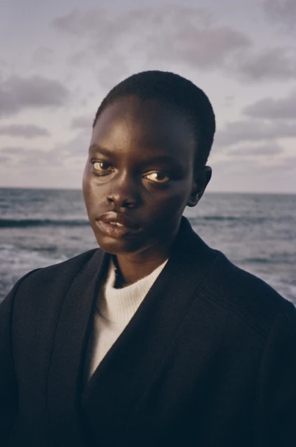 Close-up portrait of a dark-skinned adult woman with short hair, wearing a dark coat, with the ocean and sky visible in the background.